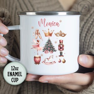 May include: White enamel mug with a pink and gold Christmas design. The mug has the name "Monica" written on it. The design includes a ballerina, a nutcracker, a rocking horse, a Christmas tree, and a crown. The mug is 12 ounces.