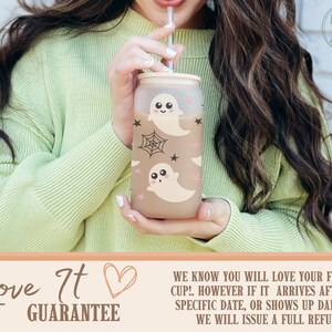 May include: Frosted glass cup with a bamboo lid and straw, decorated with cute cartoon ghosts, spiderwebs, and stars. The cup is held by a person wearing a light green sweater. The cup has the text "Love It Guarantee".