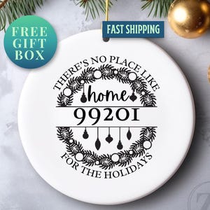 May include: White ceramic Christmas ornament with the text "There's no place like home for the holidays" and the postcode 99201. The design includes a wreath and hanging ornaments. A "Free Gift Box" sticker is visible.