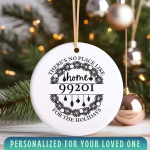 May include: White ceramic Christmas ornament with gold hanging ribbon. The ornament has the text "There's no place like home 99201 for the holidays" in black script. The bottom of the ornament reads "Personalized for your loved one".