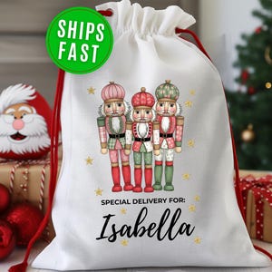 May include: White drawstring gift bag featuring three nutcracker figures in pink, red, and green outfits. The bag has red drawstrings and the text "Special Delivery For: Isabella" in black. A green circle with "SHIPS FAST" is at the top.