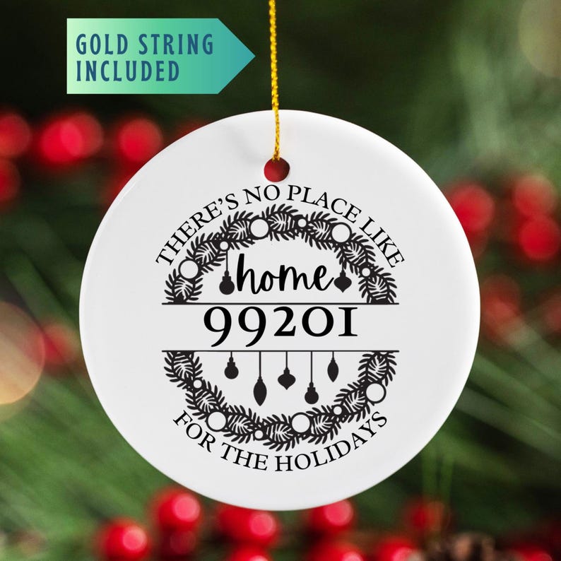 May include: A white ceramic holiday ornament with the text "THERE'S NO PLACE LIKE home FOR THE HOLIDAYS" and the zip code 99201. The design includes a wreath and hanging ornaments. A gold string is attached for hanging.