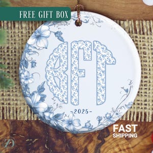 May include: A round, white ceramic ornament with a blue floral design and the initials "RFT" in a floral pattern. The year "-2025-" is printed below the initials. The ornament has a gold string for hanging. The words "FREE GIFT BOX" are printed on a green rectangle.