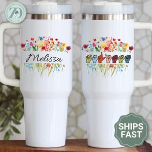 May include: White travel mug with a handle, featuring a floral design with the name "Melissa" and a sign language alphabet. The mug has a silver rim and a white lid. The handle is white.