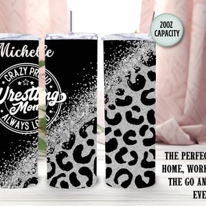 May include: A black and silver tumbler with a lid and straw. The tumbler features the text "Michelle Crazy Proud Wrestling Mom Always Loves" in white. The design includes a leopard print pattern and a 591ml capacity.