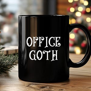 May include: Black ceramic mug with the white text "OFFICE GOTH". The mug has a rounded handle and a glossy finish. The mug is sitting on a wooden surface with a blurred background of Christmas lights.