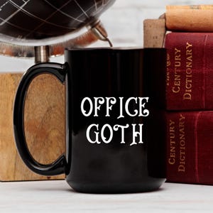 May include: Black ceramic mug with the words "OFFICE GOTH" in white, stylized font. The mug is set against a backdrop of books and a globe, suggesting a workspace or office setting.
