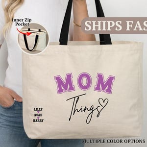 May include: Cream-colored tote bag with black handles, featuring the words "MOM Things" in purple and black script. The bag includes the names "Lilly, Noah, Harry" and a heart symbol. An inner zip pocket is visible.