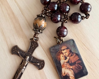 May include: A rosary featuring dark red, polished spherical beads connected by bronze wire. It includes a bronze crucifix with intricate floral engravings and a rectangular pendant depicting a figure holding a child, accented by a single tan bead.