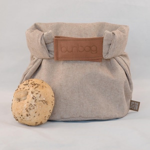Könnte beinhalten: Eine beigefarbene Brottasche aus Leinen mit einem braunen Lederriemen und dem eingeprägten Wort "bunbag". Die Tasche liegt auf einer weißen Oberfläche neben einem runden Brötchen mit Kümmelsamen.