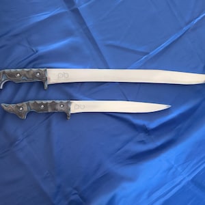 Könnte beinhalten: Zwei Messer mit silbernen Klingen und dunklen, strukturierten Griffen. Das größere Messer hat eine lange, gerade Klinge, während das kleinere eine kürzere, gebogene Klinge hat. Beide Messer haben die Initialen "pb" auf der Klinge eingraviert.