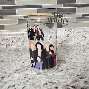 May include: A clear glass tumbler with a bamboo lid, featuring a group of stylized figures in black and other colors. The text "The Witch's Craft Room" is visible on the side. The tumbler is on a speckled countertop.
