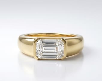 May include: A gold ring featuring a central emerald-cut clear gemstone set in a smooth, polished yellow gold band. The rectangular stone is held in a bezel setting that integrates seamlessly into the thick, rounded profile of the metal band.