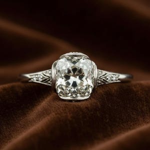 Pode incluir: Um anel de platina com um grande diamante de corte quadrado como peça central. O diamante brilha com muitas facetas, e a banda apresenta intrincados filigranas e diamantes de acento menores. O anel está sobre um fundo de veludo castanho escuro.