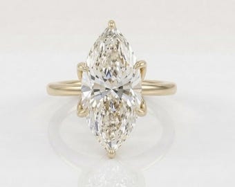 May include: A marquise-cut diamond ring set in a polished yellow gold band. The center stone is elongated with pointed ends and features multiple facets that reflect light. The setting includes six prongs that secure the diamond to the gold band.