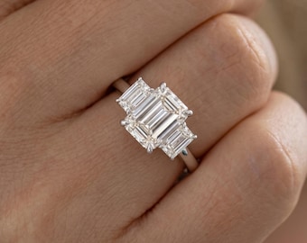 May include: A three-stone ring featuring a large emerald-cut center diamond flanked by two smaller emerald-cut diamonds. The stones are set in a polished silver-toned metal band with four prongs securing the center stone and prongs on the sides.
