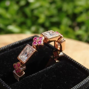 May include: A rose gold ring with a geometric design. The ring features clear, square-cut and rectangular-cut stones, along with small, round, pink stones. The ring is displayed in a black velvet box against a blurred green background.
