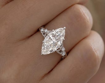 May include: A silver-toned ring featuring a large marquise-cut center stone held by prongs. The band is accented with smaller round stones on each side, integrated into a delicate, curved metal setting that frames the central gemstone.