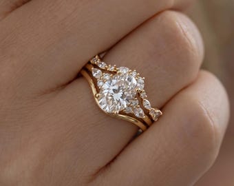 May include: A gold ring set featuring a central oval-cut white stone surrounded by smaller marquise and round-cut stones, paired with a curved gold band. The metal has a polished finish, and the stones are set in a cluster arrangement.