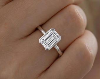 May include: A silver-toned ring featuring a large, rectangular emerald-cut clear gemstone set in a sleek, polished metal band. The stone is held by four prominent prongs at the corners, showcasing a clear, faceted surface that reflects light.