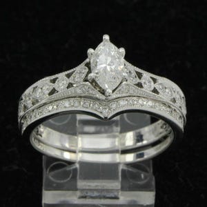May include: A marquise-cut diamond engagement ring with a matching band. The rings are silver-toned, with intricate detailing and small accent diamonds. The engagement ring features a large, central marquise-cut diamond. The band is curved to fit the engagement ring.