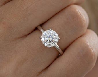 May include: A solitaire ring featuring a round brilliant-cut clear gemstone set in a polished silver-toned metal band. The stone is secured by six prongs, showcasing a faceted surface that reflects light with a bright, clear, and sparkling appearance.