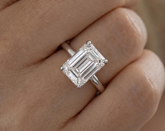May include: A silver-toned ring featuring a large, rectangular emerald-cut clear gemstone set in a four-prong setting. The band is smooth and polished, showcasing a clean, reflective surface that highlights the geometric facets of the center stone.