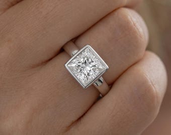 May include: A silver-toned ring featuring a square-cut, clear, faceted gemstone set in a smooth bezel setting. The band is plain and polished, showcasing a clean, metallic finish that encircles the finger with a simple, modern design.