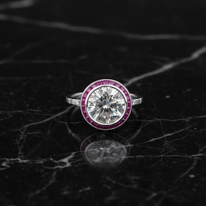 Peut inclure: Une bague en platine avec une grosse pierre précieuse ronde et transparente au centre, entourée d'un halo de petites pierres précieuses rouges. La bague est posée sur une surface en marbre noir, reflétant le design.