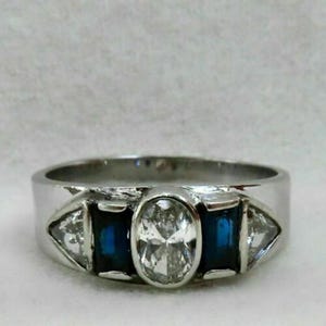May include: A silver ring with an oval-cut, clear center stone, flanked by two rectangular, dark blue stones and two triangular, clear stones. The ring has a wide band.