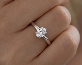 May include: A solitaire ring featuring a clear, oval-cut gemstone set in a polished silver-toned metal band. The stone is secured by four prongs, showcasing a faceted surface that reflects light, mounted on a smooth, plain band.