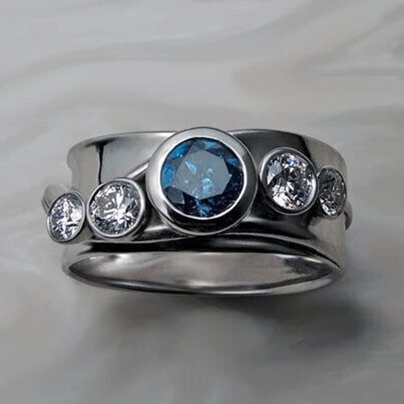 May include: A silver ring with a blue gemstone and three smaller white gemstones. The ring is made of two bands, one wider than the other.