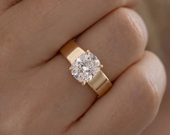May include: A gold ring featuring a wide, polished band and a single cushion-cut clear gemstone held in place by four prongs. The metal has a warm, reflective finish, and the stone is faceted to catch the light, creating a brilliant appearance.