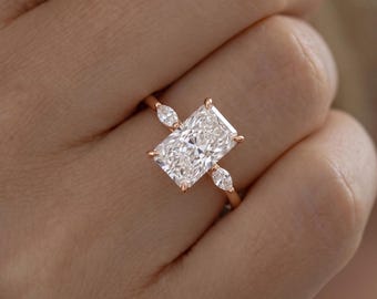 May include: A rose gold ring featuring a large rectangular radiant-cut center stone held by four prongs, flanked by two smaller marquise-cut side stones. The band is thin and polished, showcasing a clear, faceted design with brilliant light reflection.