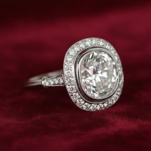 May include: A silver ring with a large, clear, cushion-cut diamond center stone. The diamond is surrounded by a halo of smaller diamonds, and the band also features diamond accents. The ring is set against a deep red background.