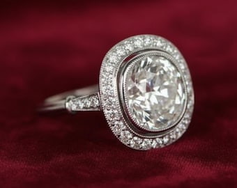 Könnte beinhalten: Ein silberner Ring mit einem großen, klaren Diamanten im Kissenschliff als Mittelstein. Der Diamant ist von einem Halo aus kleineren Diamanten umgeben, und auch das Band weist Diamantakzente auf. Der Ring ist vor einem tiefroten Hintergrund platziert.