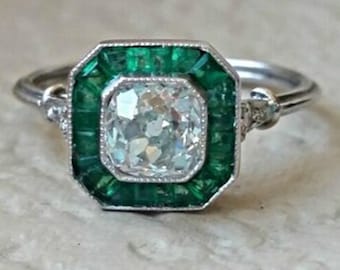 Könnte beinhalten: Ein silberner Ring mit einem quadratisch geschliffenen, klaren Edelstein in der Mitte, umgeben von einem Rand aus smaragdgrünen Steinen. Der Ring hat ein detailliertes Design am Band und an der Fassung, was einen Vintage- oder Antik-Stil suggeriert.