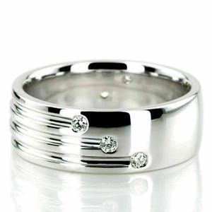 May include: A polished silver ring with three small diamonds set in a row. The ring has a wide band with horizontal grooves on one side. The diamonds are round and sparkle against the smooth, reflective metal.