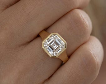 May include: A gold ring featuring a large, square-cut, clear gemstone set in a bezel setting. The band is smooth and solid gold, with the stone positioned centrally in an octagonal frame that highlights the faceted clarity of the center gem.