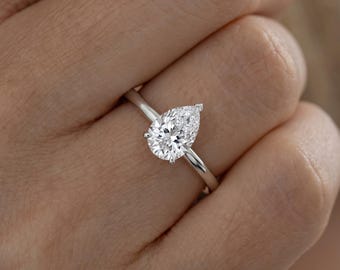 May include: A pear-shaped, brilliant-cut clear gemstone set in a polished silver-toned metal band. The stone is secured by a multi-prong setting, showcasing a faceted surface that reflects light. The ring features a slender, smooth, and rounded band.