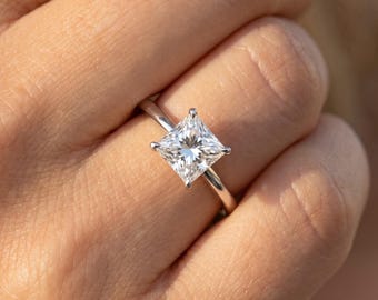 May include: A solitaire ring featuring a princess-cut clear gemstone set in a polished silver-toned metal band. The square-shaped stone is held in place by four prongs, showcasing a faceted surface that reflects light from multiple angles.