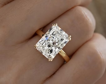 May include: A radiant-cut clear gemstone ring set in a polished yellow gold band. The large rectangular stone features multiple facets that reflect light, secured by four small gold prongs at each corner of the setting on a smooth gold band.