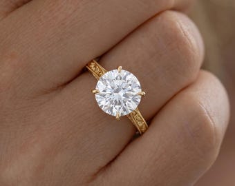 May include: A gold ring featuring a large, round, brilliant-cut clear gemstone held by four prongs. The band is crafted from yellow gold and displays intricate, embossed floral or scrollwork detailing along the shank on both sides.