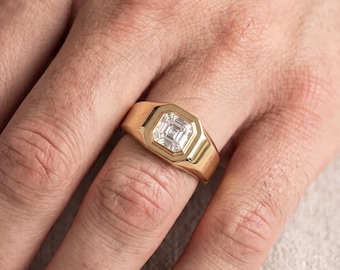 May include: A gold signet ring featuring a square-cut center stone with a step-cut facet pattern. The ring has a polished, smooth band that tapers slightly toward the back, showcasing a clean, metallic finish and a clear, faceted gemstone.