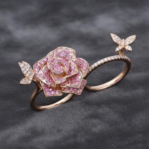 May include: A rose gold ring featuring a large pink gemstone rose and two butterfly accents. The ring has a double-finger design, with one band adorned with small white gemstones. The butterflies and rose are detailed with sparkling gemstones.