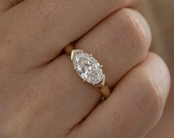 May include: A gold ring featuring a single oval-cut clear gemstone set horizontally. The stone is secured by four prongs, with two triangular gold accents flanking the center stone on the band. The band is a smooth, polished yellow gold metal.