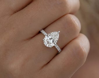 May include: A silver-toned ring featuring a large pear-shaped center stone and a band encrusted with small, clear, round-cut stones. The setting is metallic and polished, showcasing a brilliant, faceted design that reflects light across the surface.