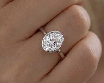 May include: A silver-toned ring featuring a large, oval-cut clear gemstone set in a smooth, polished bezel setting. The band is slender and metallic, encircling the finger with a clean, continuous design that highlights the faceted stone.