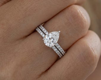 May include: A silver-toned engagement ring set featuring a pear-shaped center stone and a matching band, both adorned with small pavé-set stones along the shank. The metal band has a polished finish and the stones are clear and faceted.