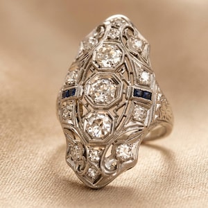 May include: An antique silver ring with a detailed filigree design. The ring features several large, clear, round-cut diamonds and smaller accent diamonds. Two small, dark blue stones are set on the ring's face. The ring is set on a beige fabric.